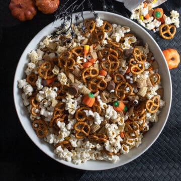 A white bowl contains a mix of popcorn, pretzels, cereal, and candy and sits on a black surface with small pumpkins.