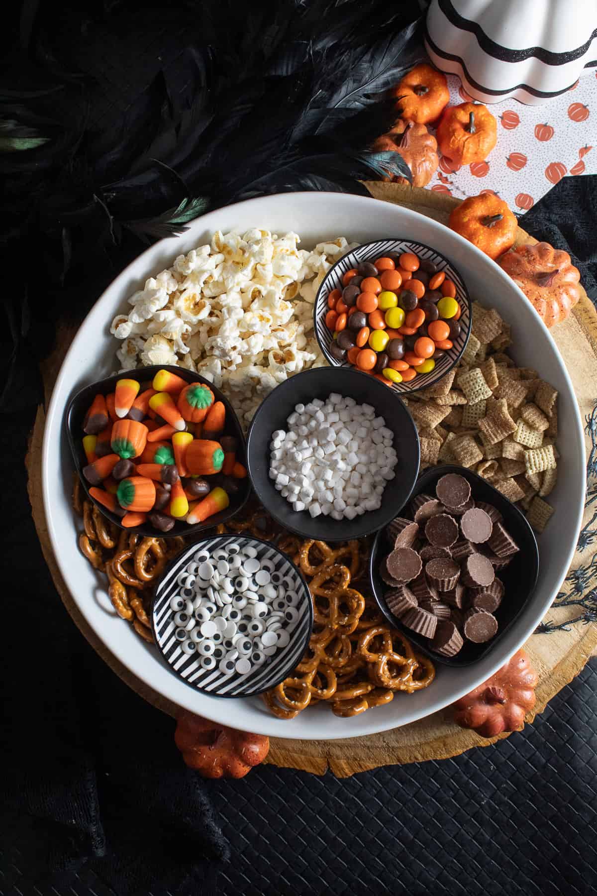 The ingredients for the snack mix are in small bowls placed in a large white platter on a black surface surrounded by pumpkins.