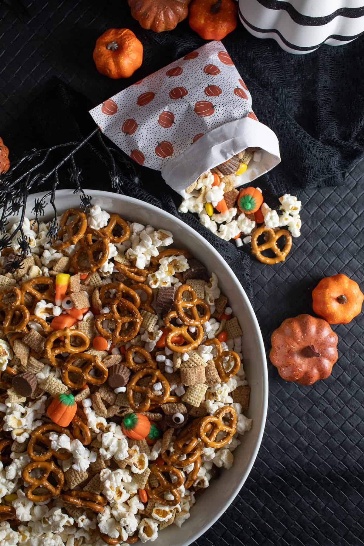 A white bowl contains a mix of popcorn, pretzels, cereal, and candy and sits on a black surface with small pumpkins and a small bag of the mixture.