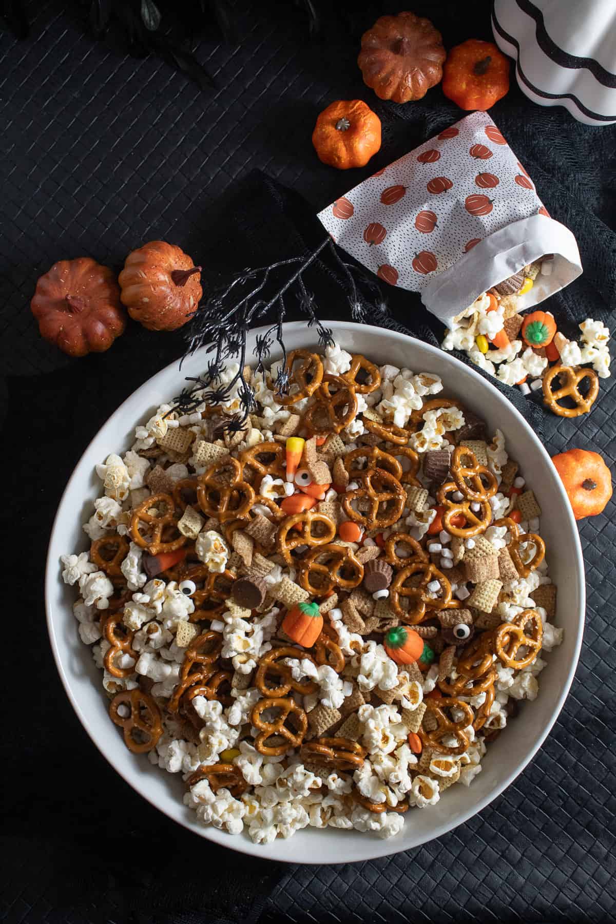 A white bowl contains a mix of popcorn, pretzels, cereal, and candy and sits on a black surface with small pumpkins and a small bag of the mixture.