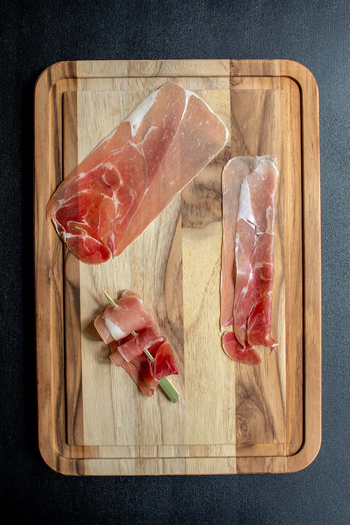 Steps for creating a prosciutto ribbon on a skewer are illustrated on a wood cutting board.