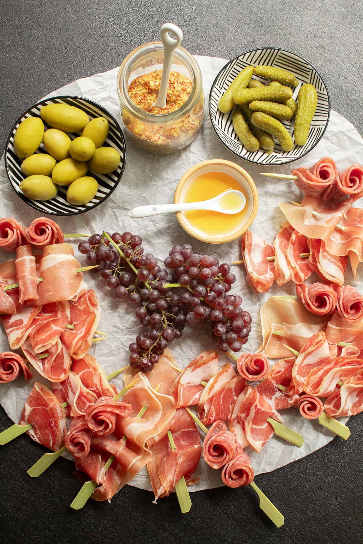 The skewers are arranged on a circle of wrinkled parchment on a black surface along with olives, mustard, honey, pickles, and grapes.