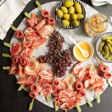 The skewers are arranged on a circle of wrinkled parchment on a black surface along with olives, grapes, mustard, gherkins, and honey.
