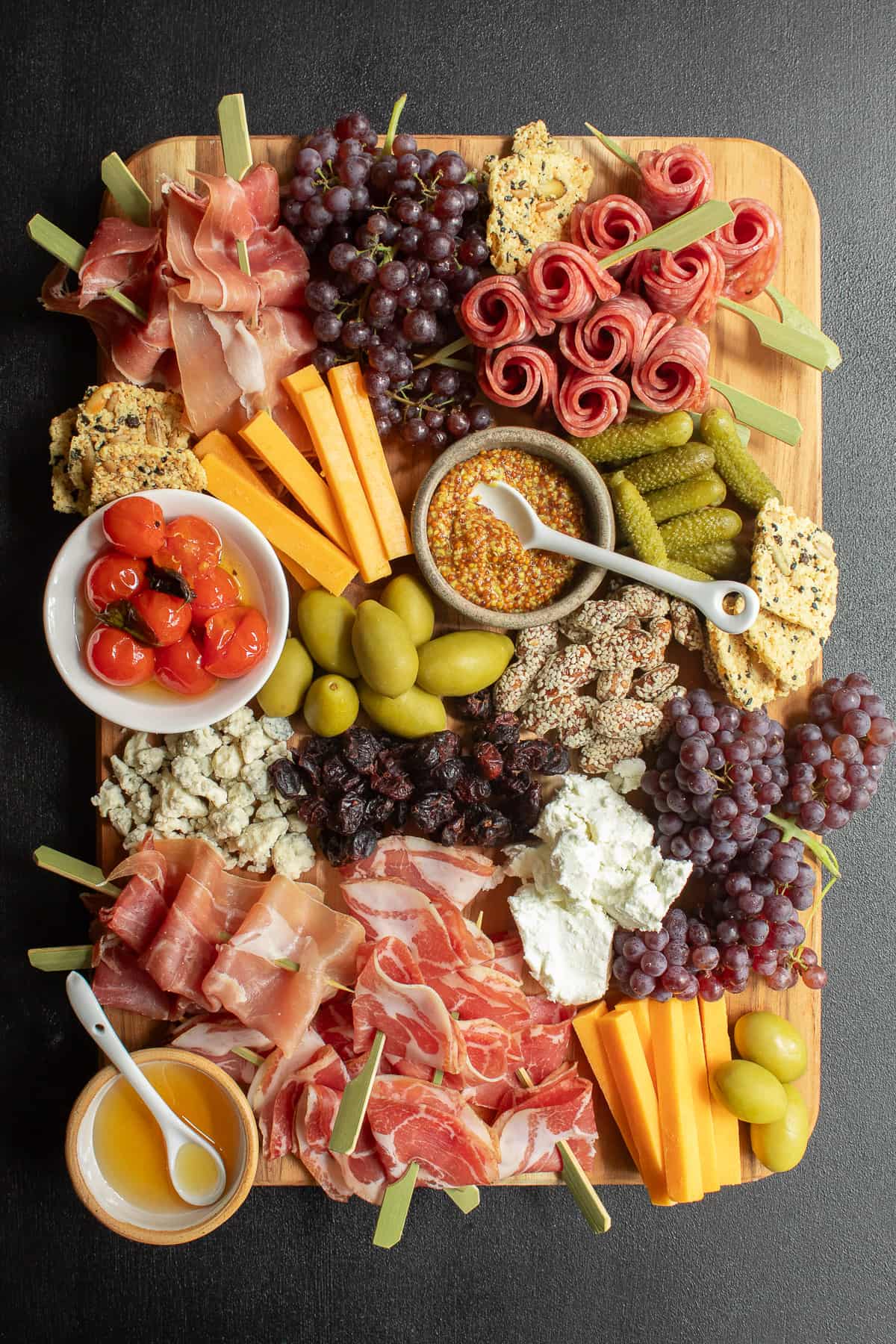 A grazing board with charcuterie, cheeses, nuts, fruit, olives, and tomatoes sits on a black surface.