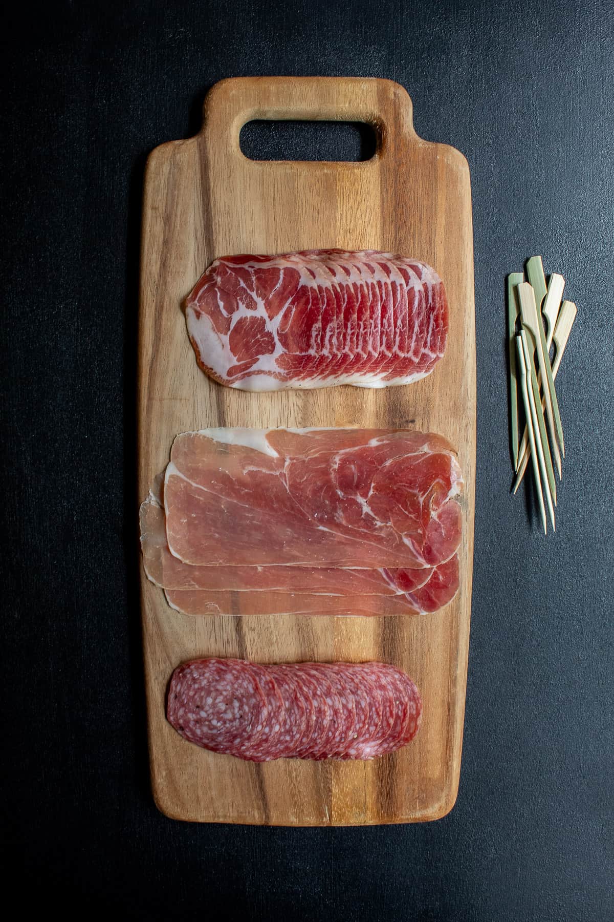 The three types of charcuterie used in the recipe are arranged on a long wooden board alongside a pile of wooden skewers.