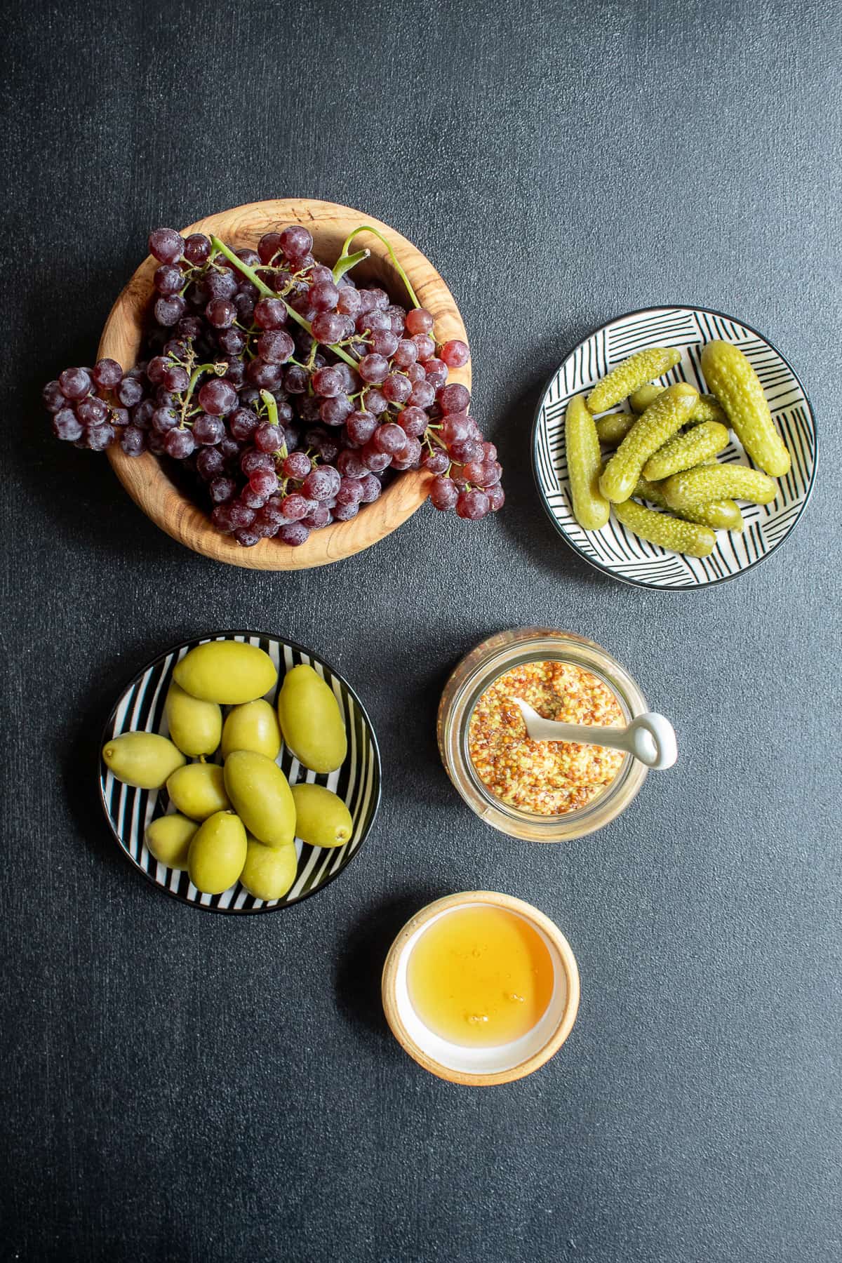 Small bowls of ingredients that compliment charcuterie: grapes, gherkins, olives, grainy mustard, and honey.