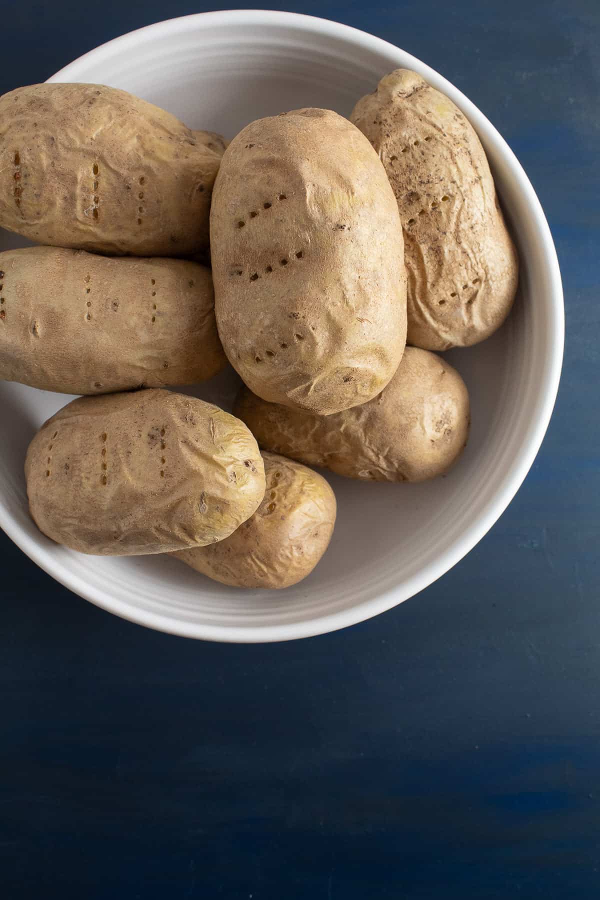 The baked potatoes are cooling in a white bowl on a blue surface.