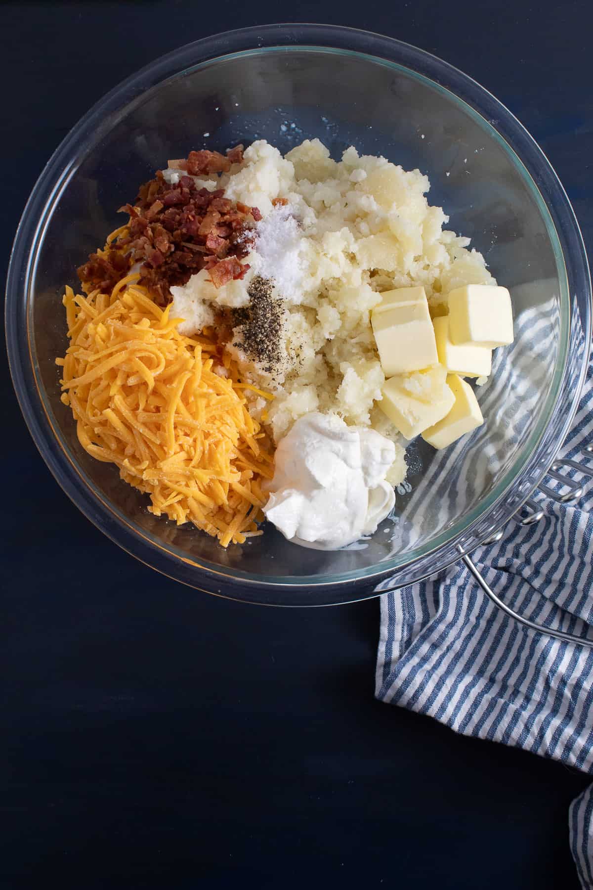 The scooped out baked potato is in a large glass bowl along with the butter, cheese, sour cream, bacon, salt, and pepper.