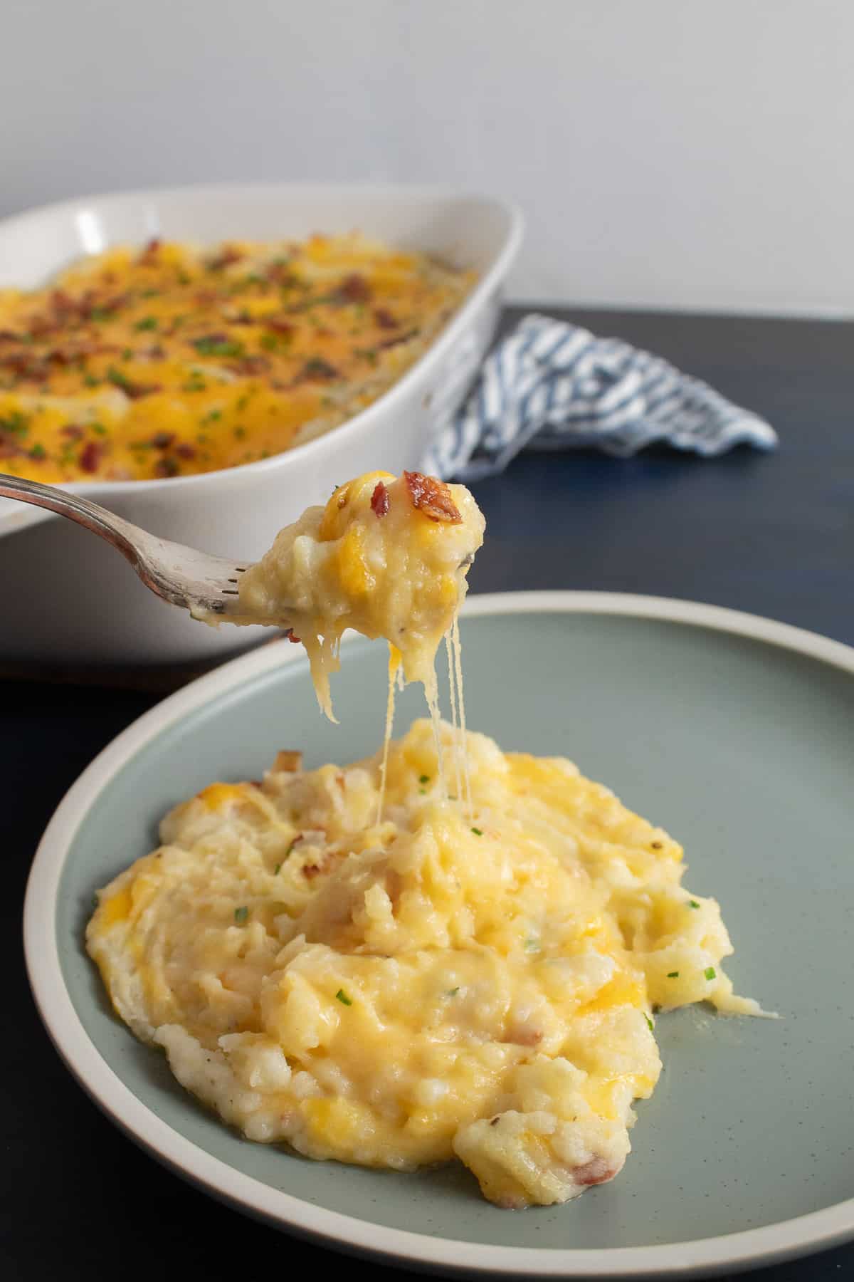 A serving of potatoes on a light blue plate with a forkful of the casserole leaving threads of cheddar cheese.