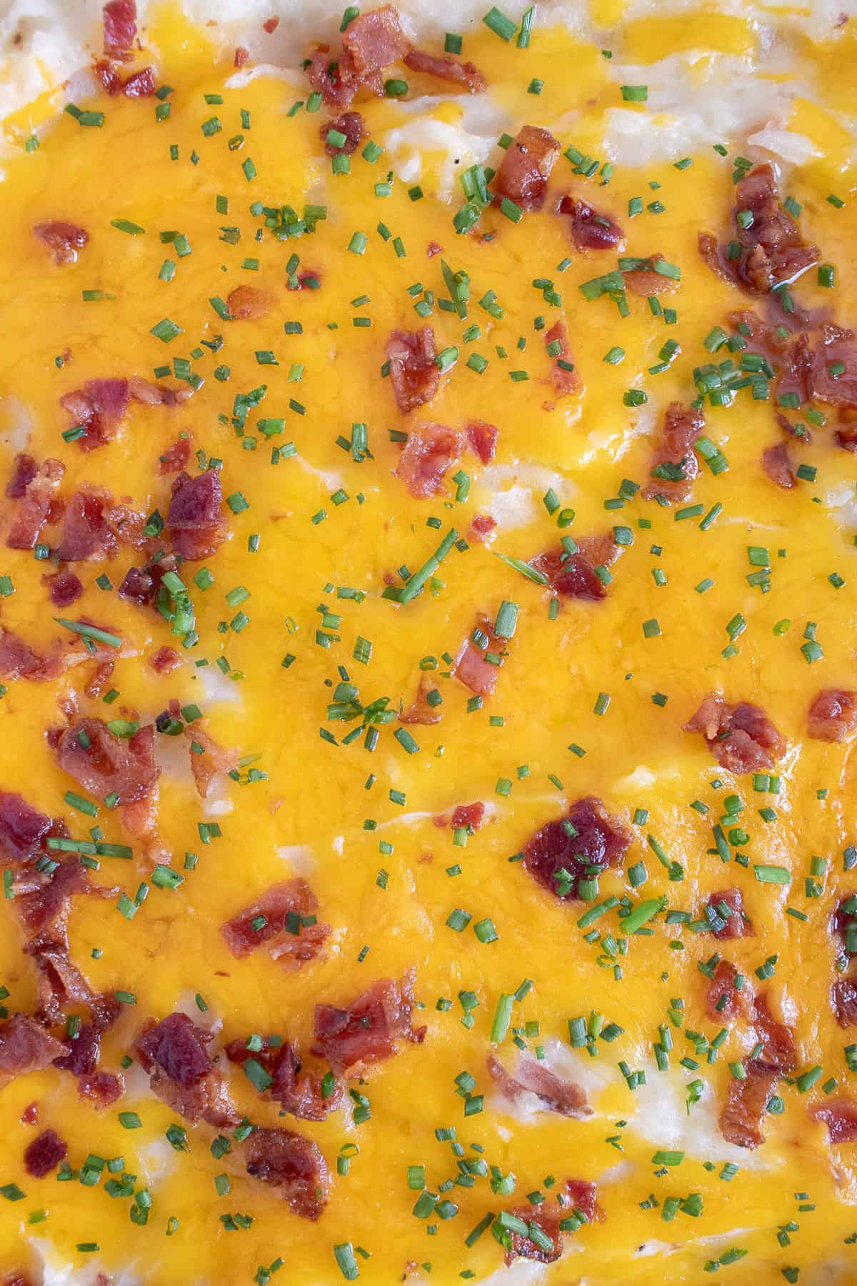 A detail image of the surface of the baked casserole with melted cheddar, finely chopped chives, and bacon pieces.