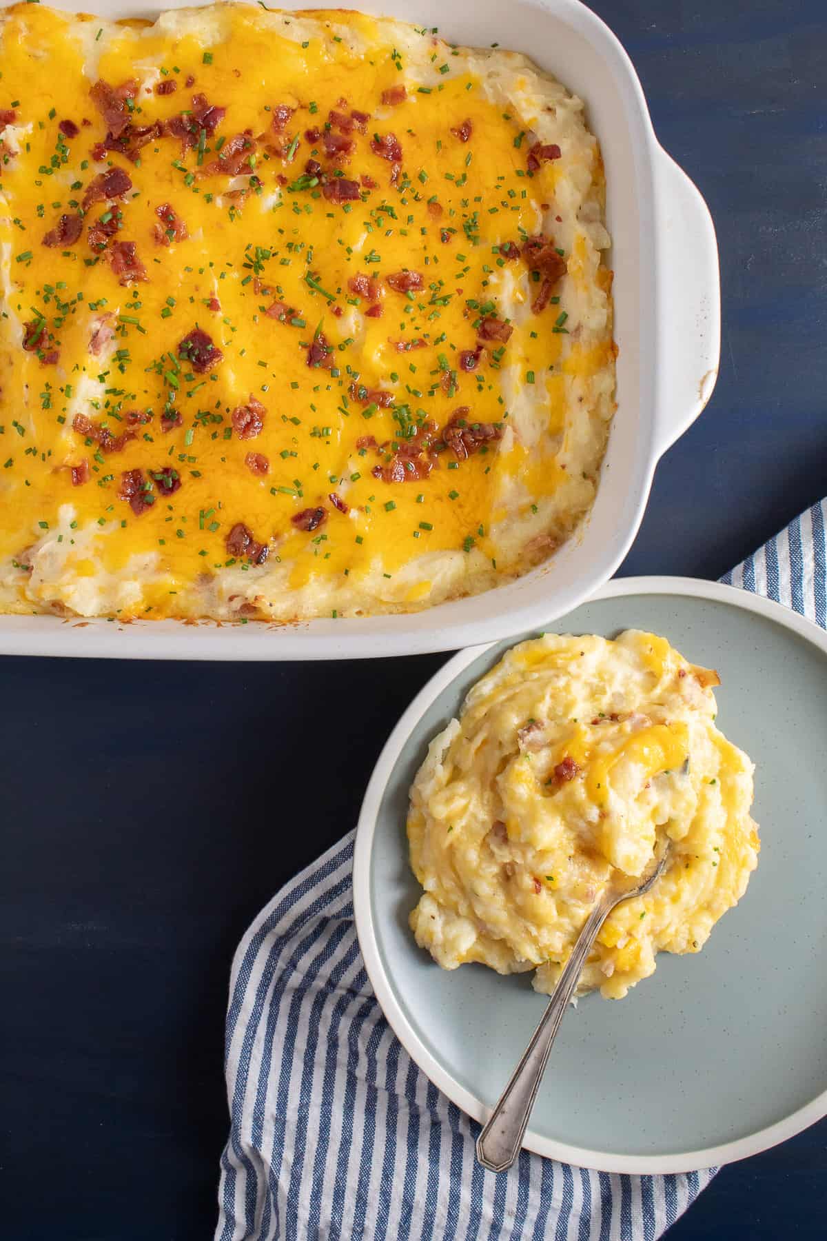 A serving of the loaded potatoes on a light blue plate sits next to a casserole dish of the mashed potatoes.