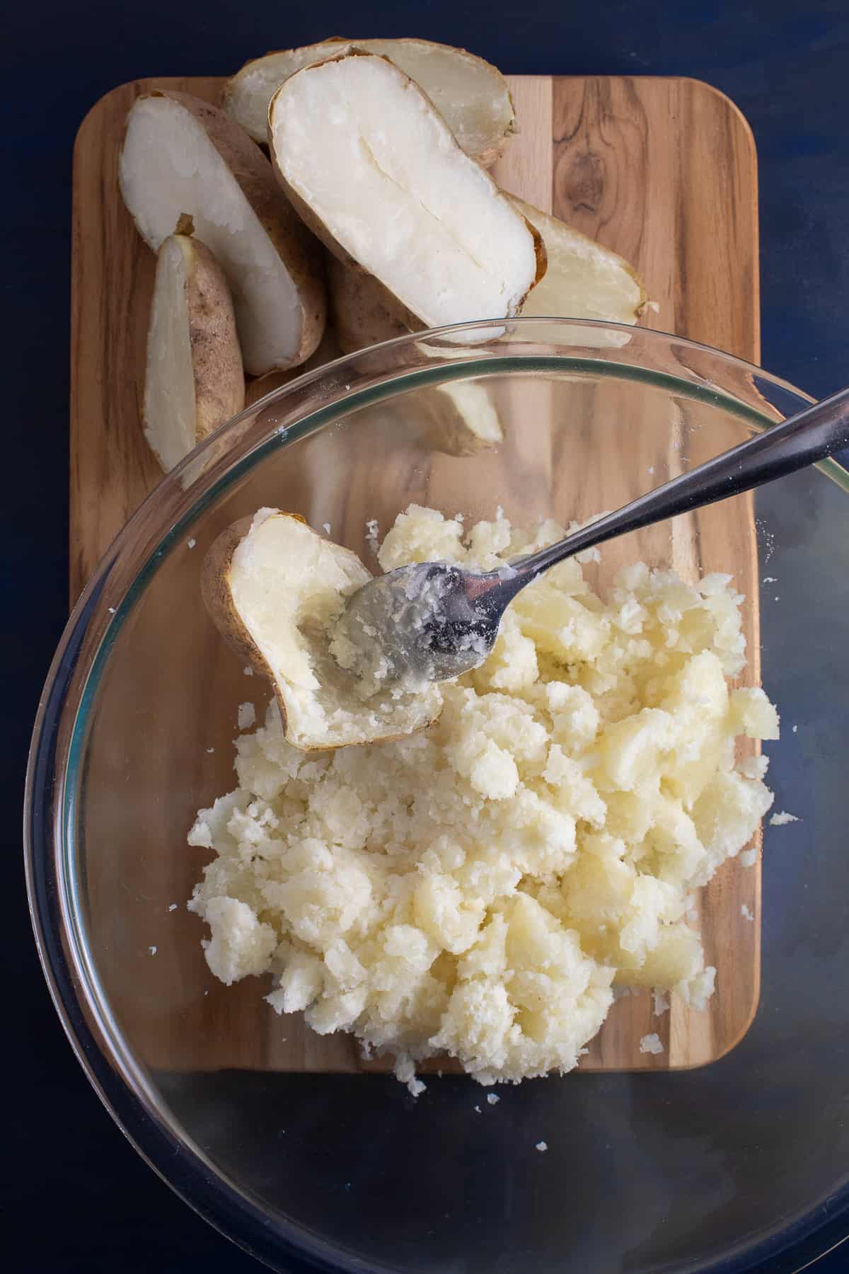 The insides of the baked potatoes are being scooped out into a large glass bowl using a spoon.