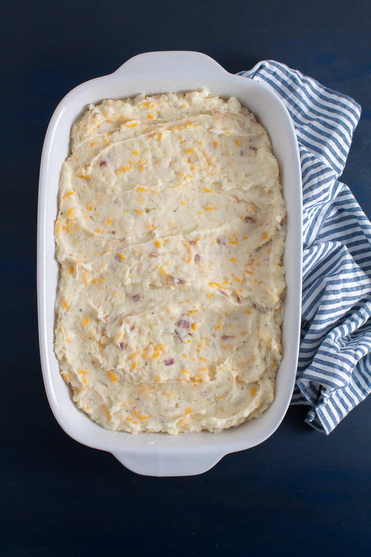 The potato mixture is spread out in a white casserole dish on a blue surface.