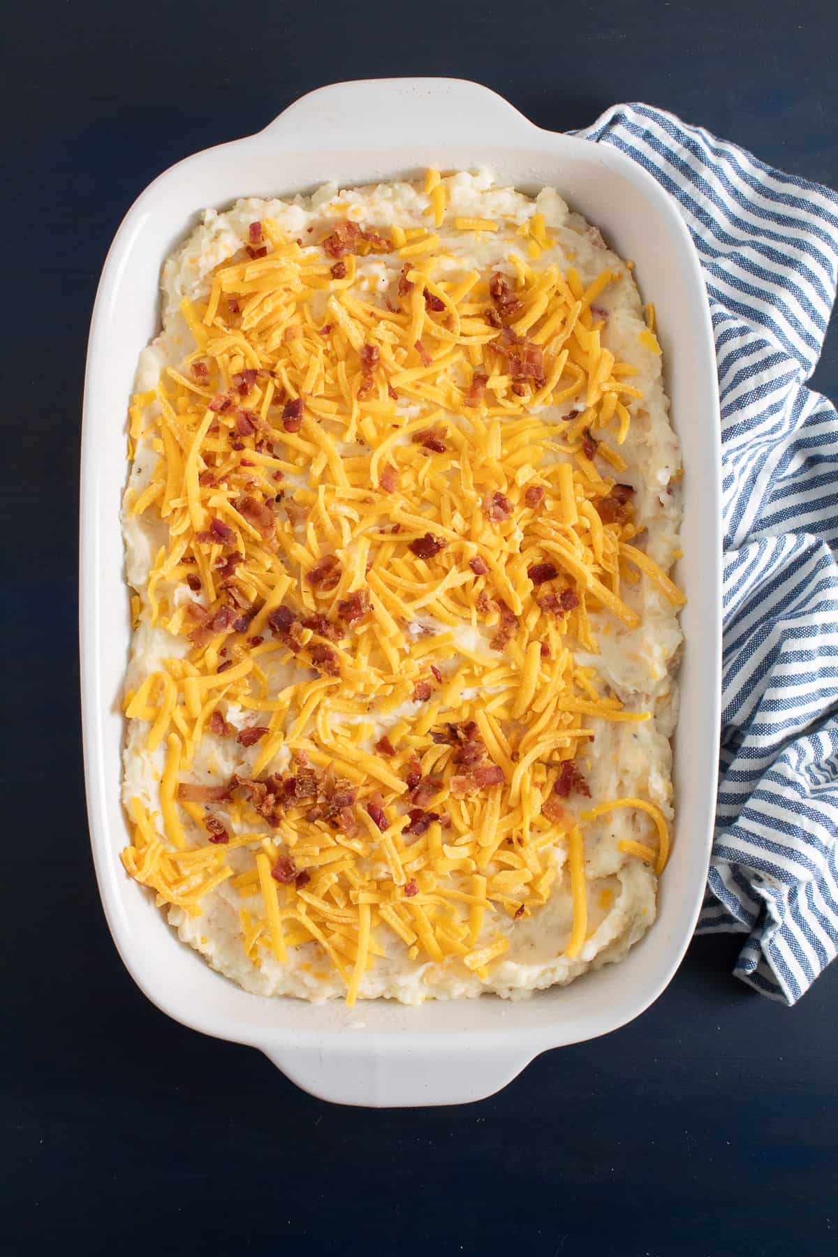 Shredded cheese and bacon pieces are sprinkled on the top of the potato mixture in a white casserole dish on a blue surface.