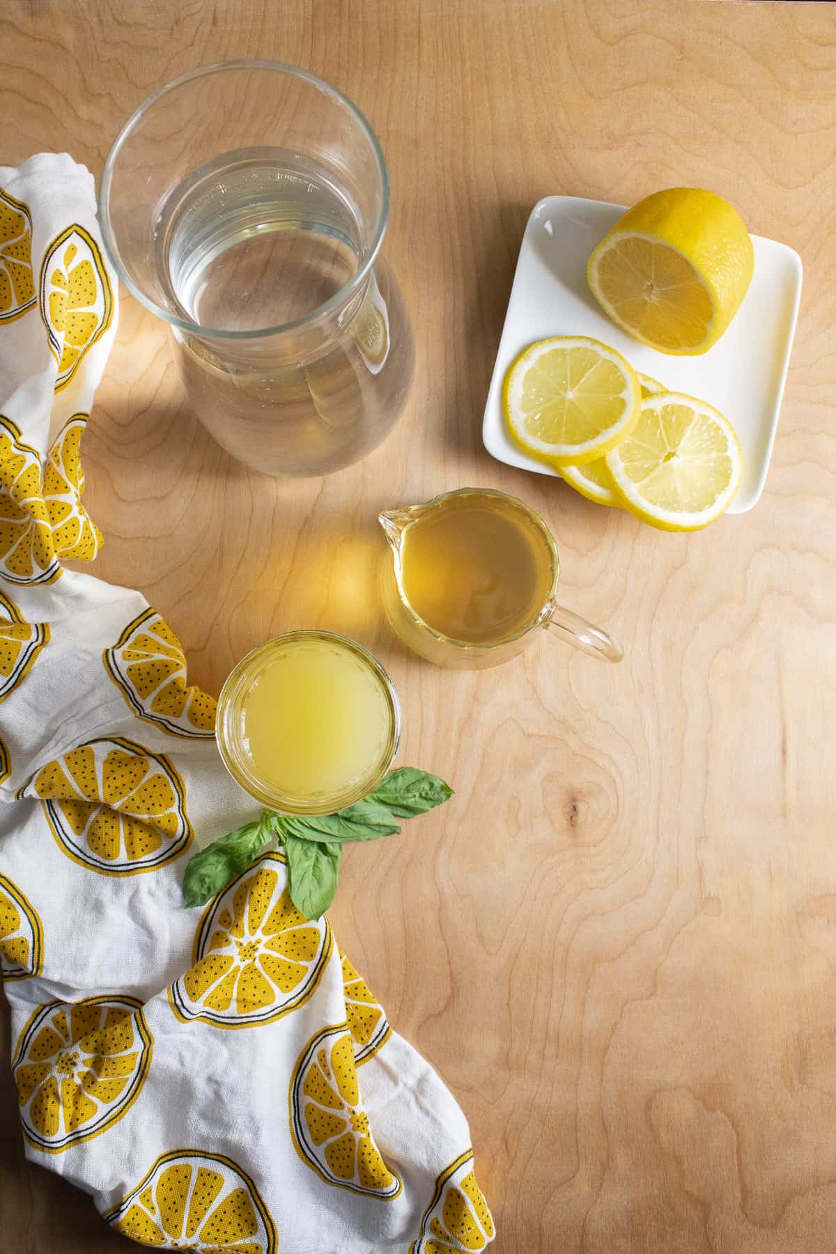 A carafe of water, fresh lemon juice, basil simple syrup, and sliced lemons are arranged on a light wooden surface.