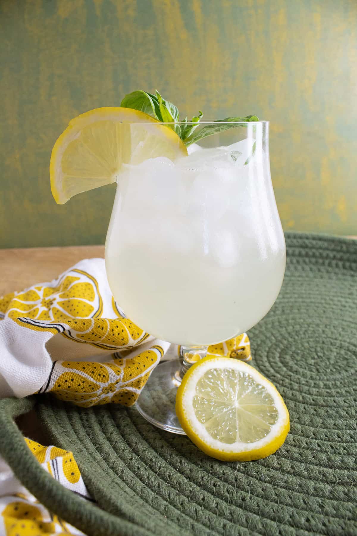 A footed glass of pale yellow lemonade is garnished with a half slice of lemon and a sprig of basil leaves.