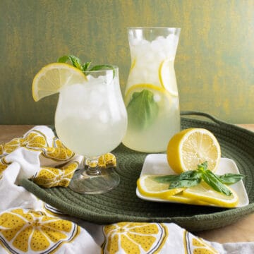 A carafe of lemonade sits on a green woven tray next to a glass of lemonade and a plate of lemon slices and basil leaves.