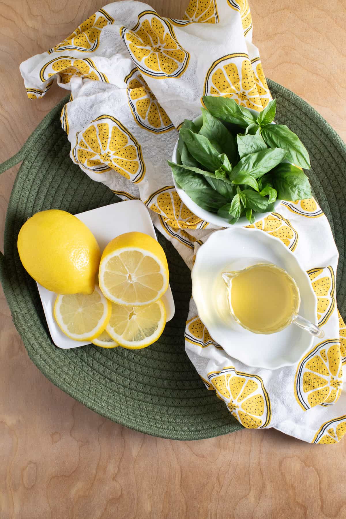 Basil leaves, fresh sliced lemons, and basil simple syrup are arranged on a green woven platter.