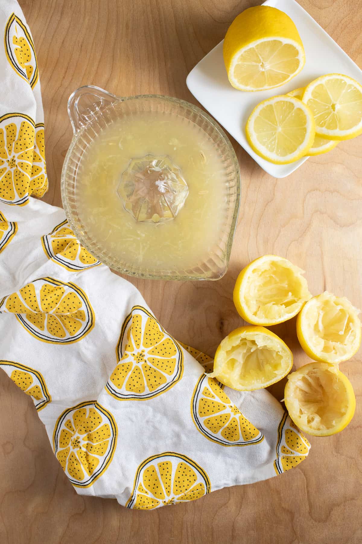 A glass juicer is full of juice and pulp and sits on a light wooden surface alongside sliced lemons and a napkin printed with lemon slices.