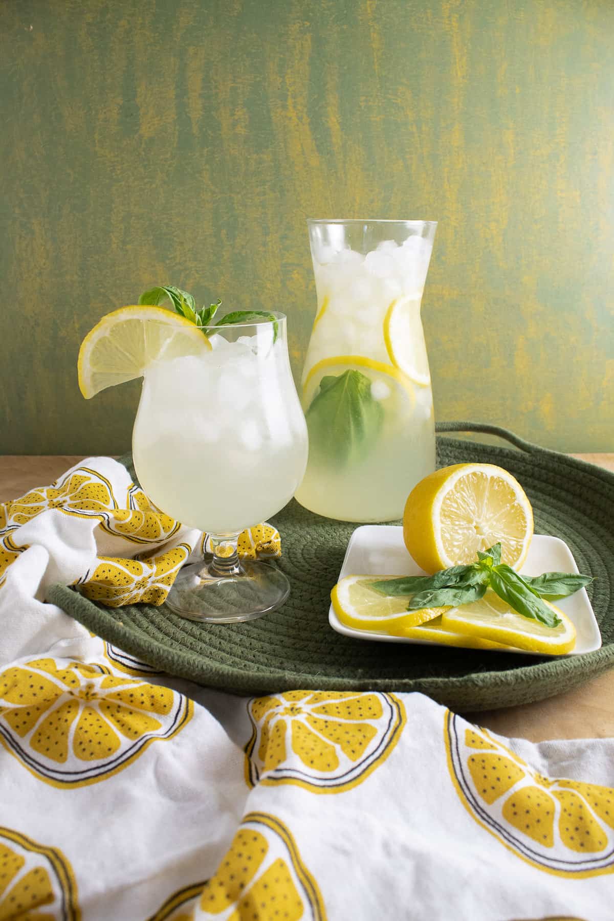 A carafe of lemonade sits on a green woven tray next to a glass of lemonade and a plate of lemon slices and basil leaves. a napkin with printed lemon slices is draped in the foreground.