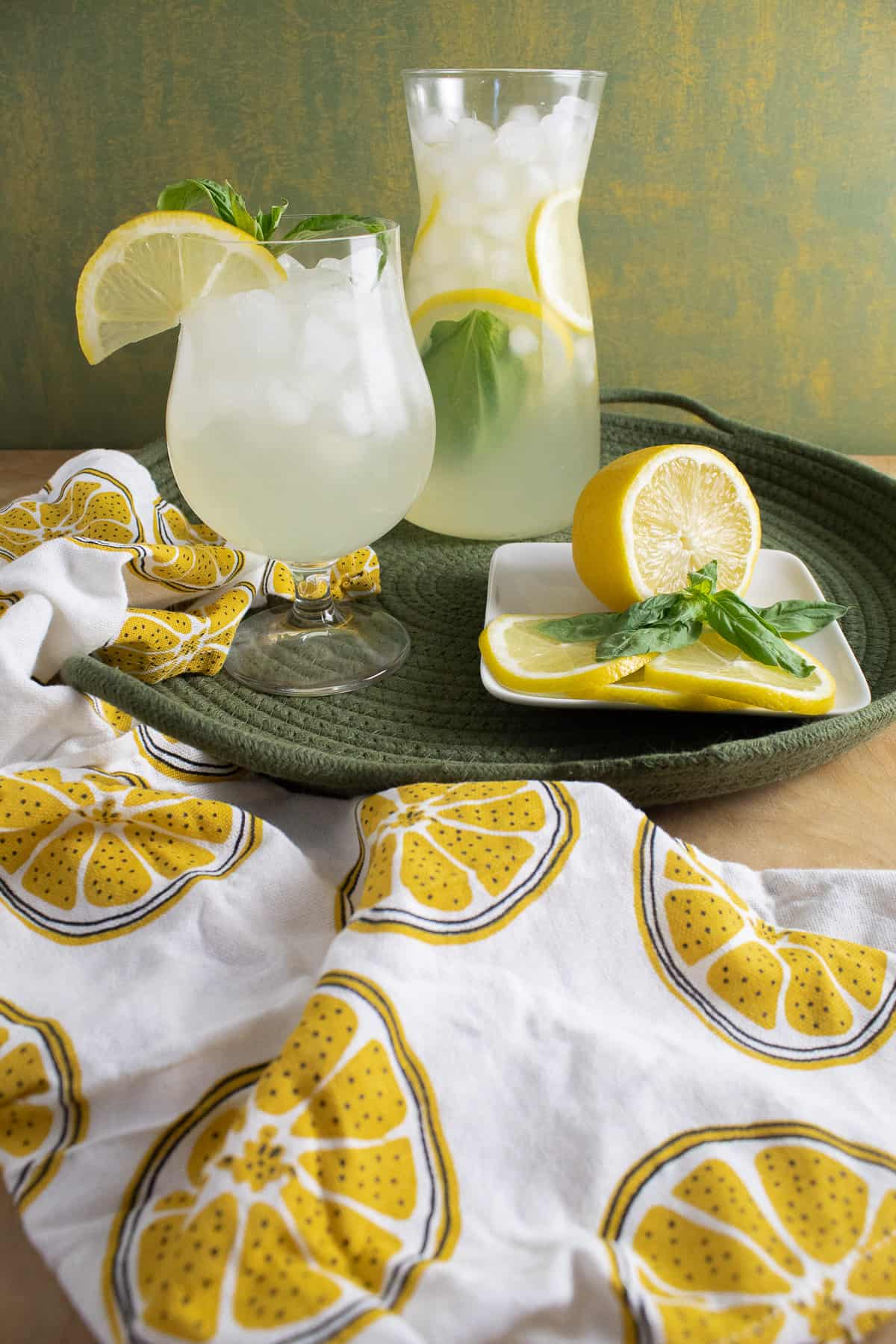 A carafe of lemonade sits on a green woven tray next to a glass of lemonade and a plate of lemon slices and basil leaves.