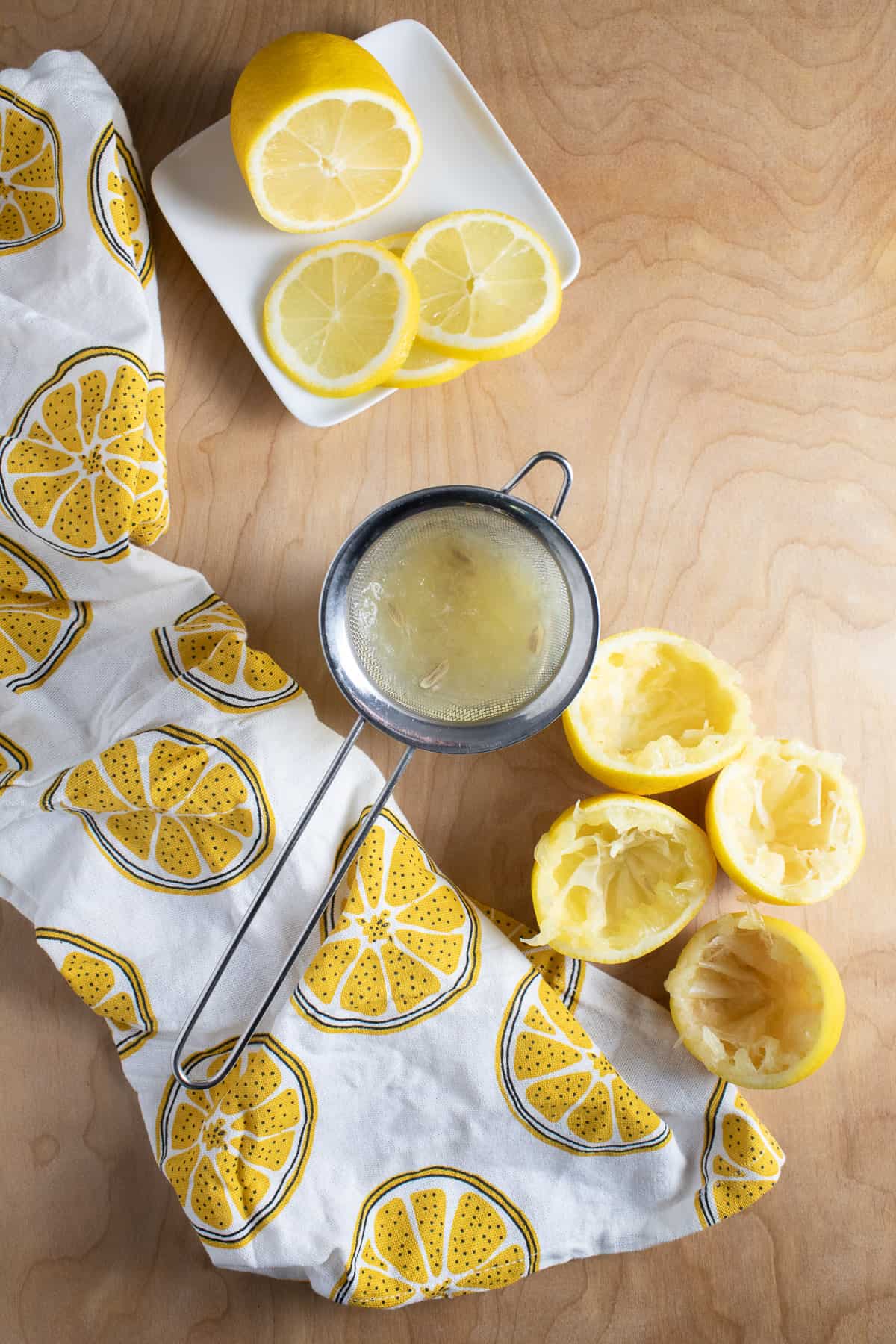 Fresh lemons are sliced in wheels or in half and sit on a light wooden surface next to a mesh strainer full of the seeds and pulp from juicing.