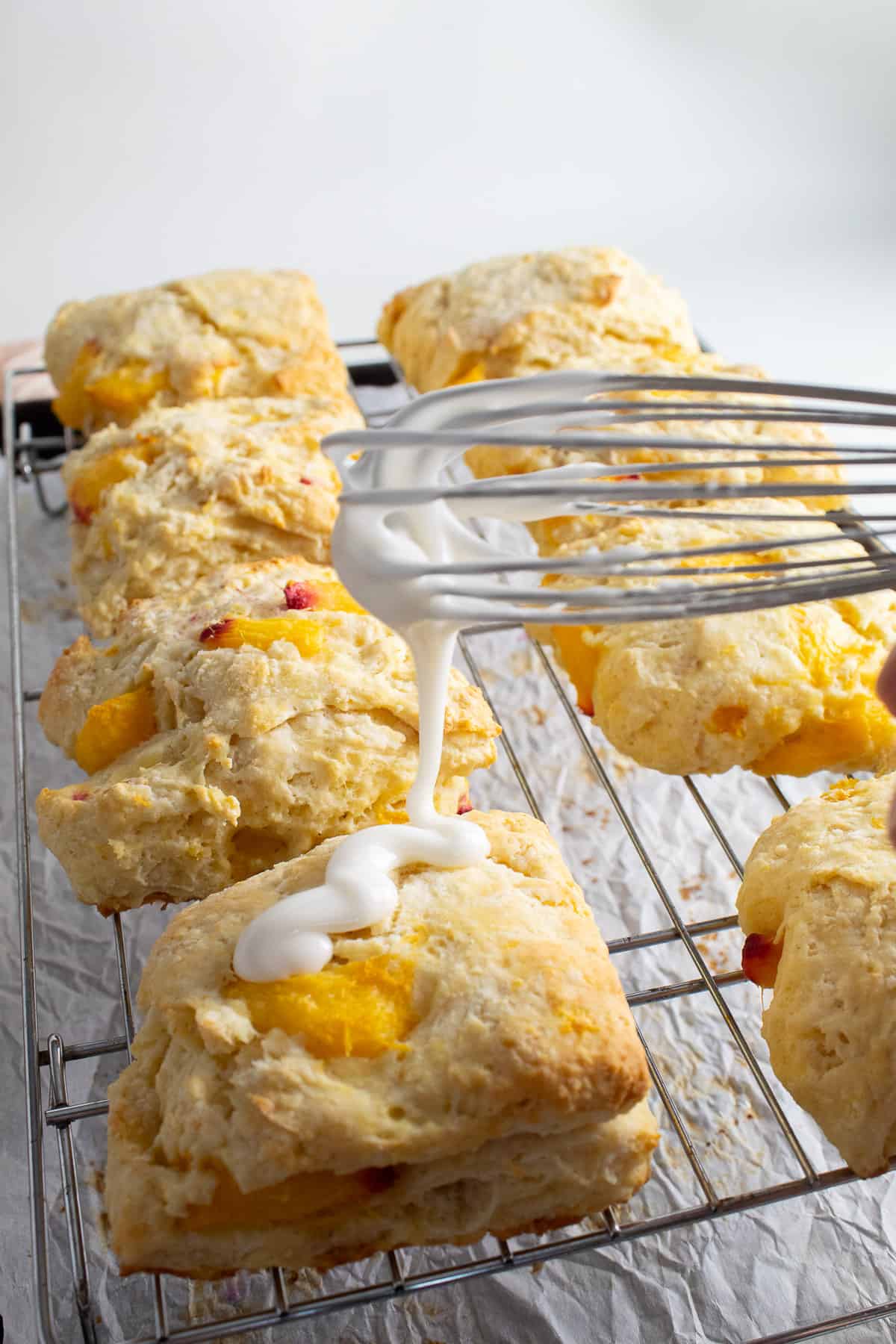 A thick white glaze is drizzled directly from the whisk onto the biscuits, which sit on a wire rack over a parchment-lined pan.