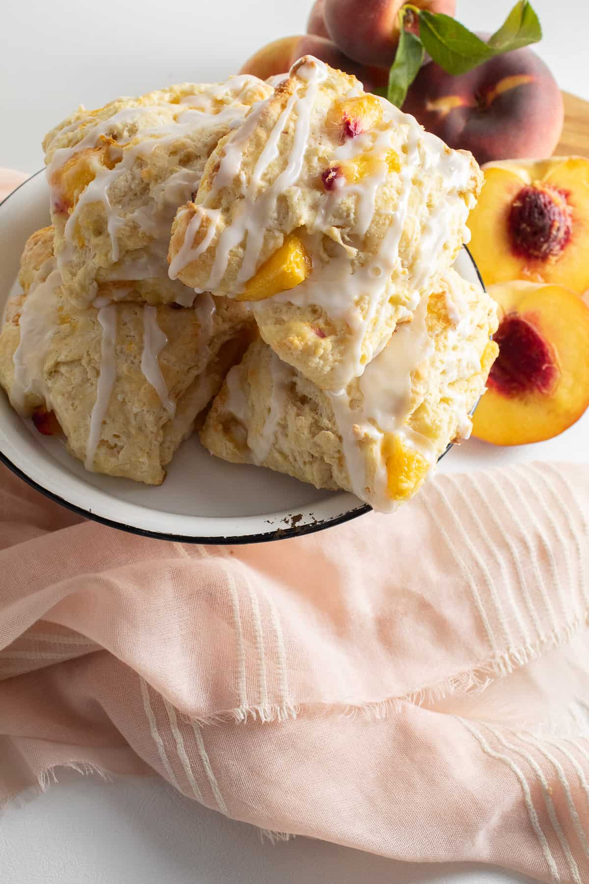 A plate of peach biscuits sits on a surface alongside a peach colored cloth and fresh halved peaches.