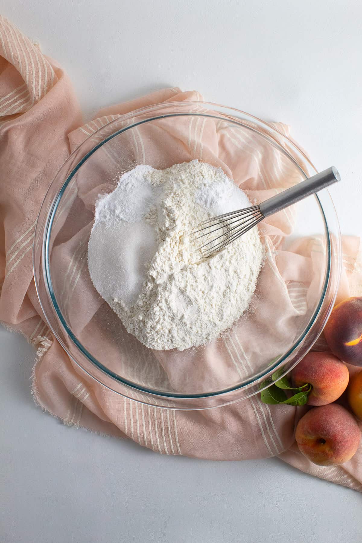 Flour, sugar, baking powder, cream of tartar, and salt are ready to be whisked together in the bottom of a large glass bowl on a white surface with a peach colored cloth.