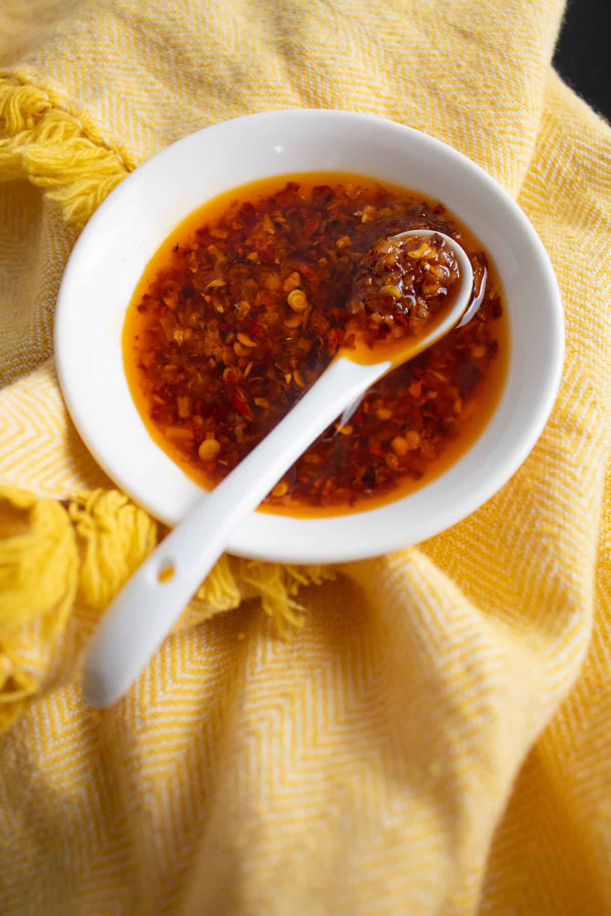 A detail image of the texture of the chili onion crunch in a white dish, with small flecks of chili surrounded by a reddish orange oil.