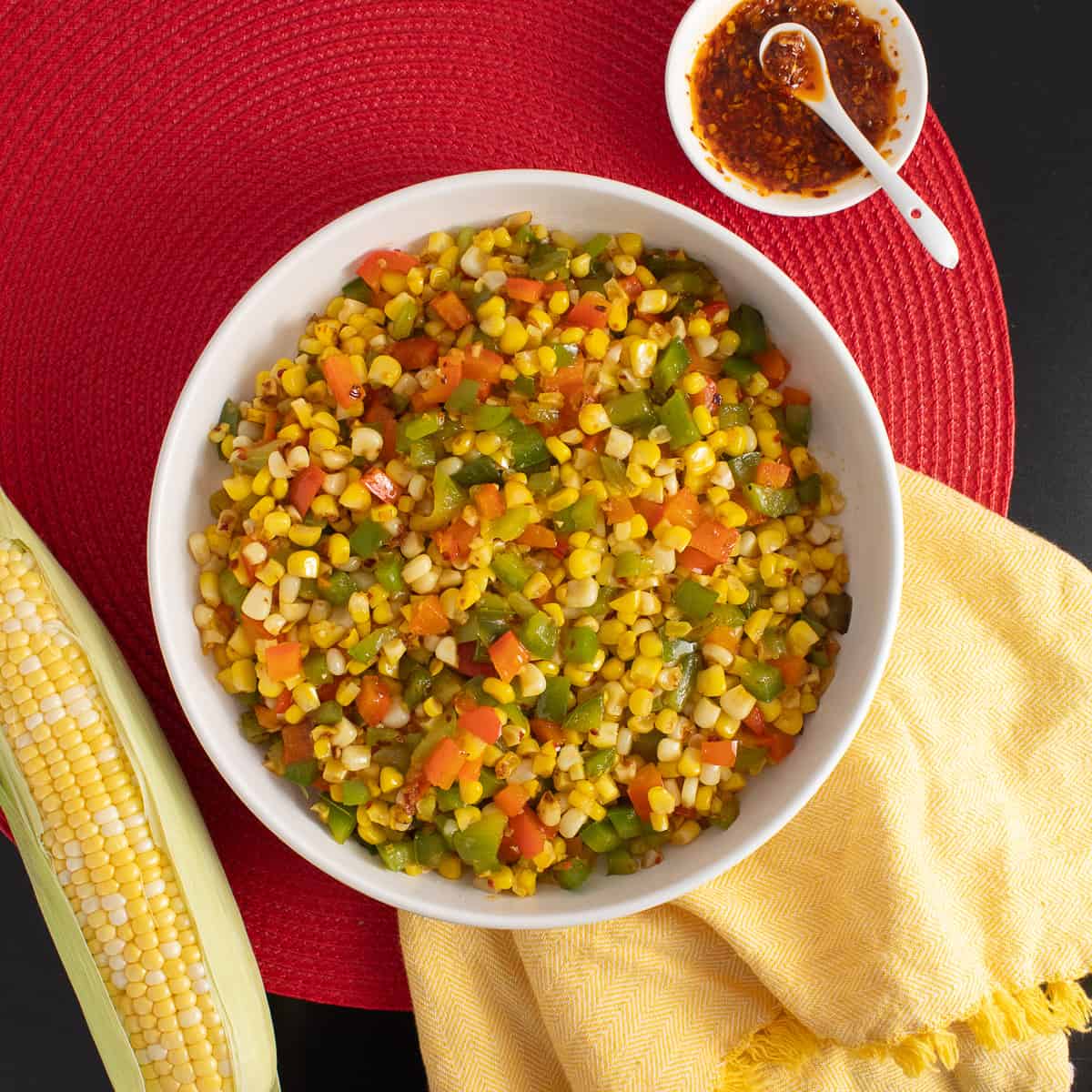 A platter of the finished dish sits on a red and black surface next to a small dish of chili onion crunch and an ear of corn.