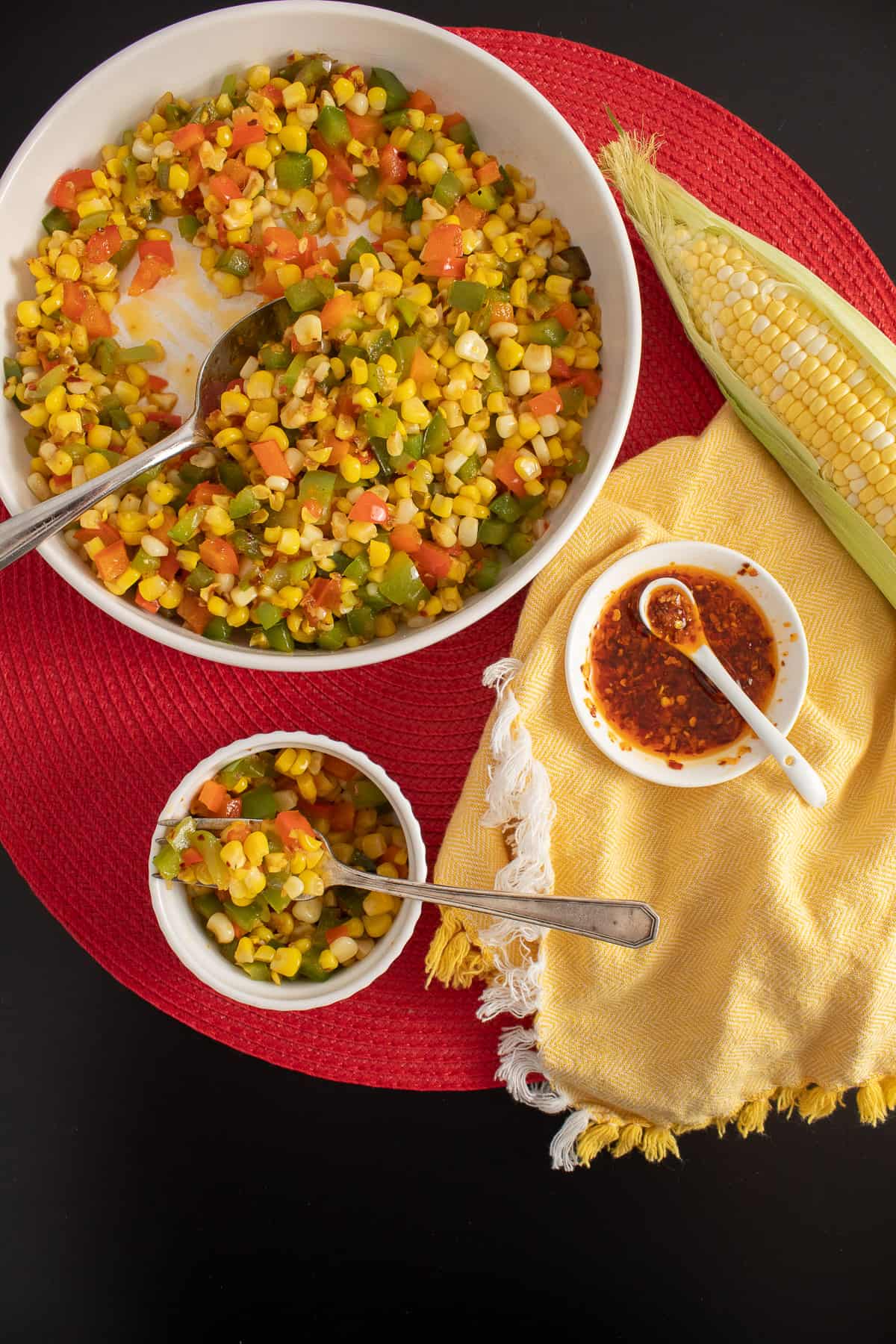 A platter of the finished dish sits on a red and black surface next to a serving in a small bowl and a small dish of chili onion crunch.