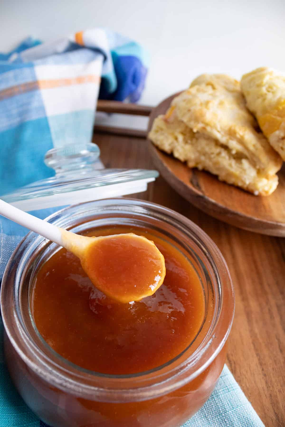 A close up detail of the smooth texture of the peach butter in a glass jar with a plate of biscuits in the background.