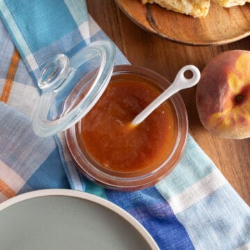 A jar of peach butter sits on a wood tray along with a fresh peach and a blue plaid cloth.