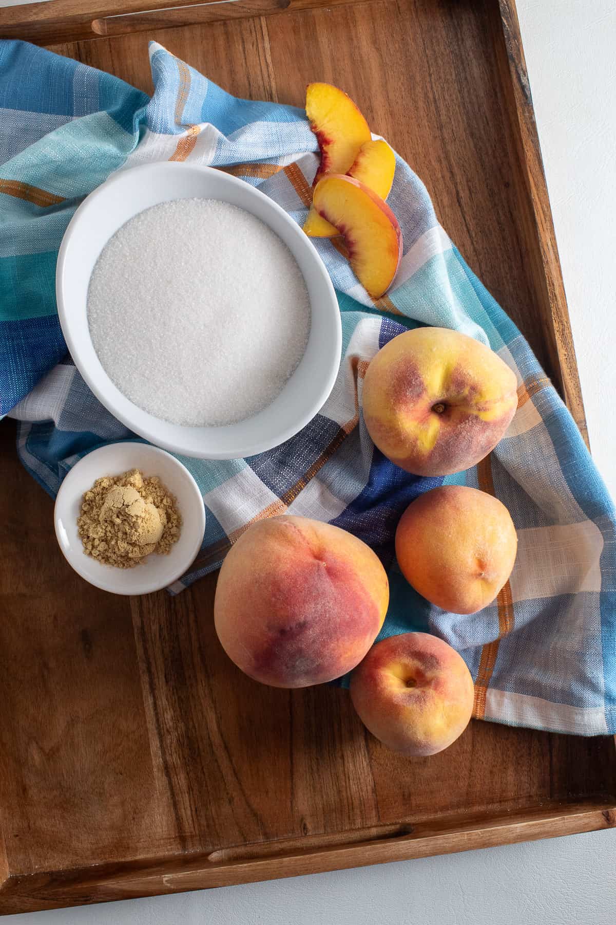 The ingredients displayed on a wooden tray: fresh peaches, granulated sugar, and ground ginger.