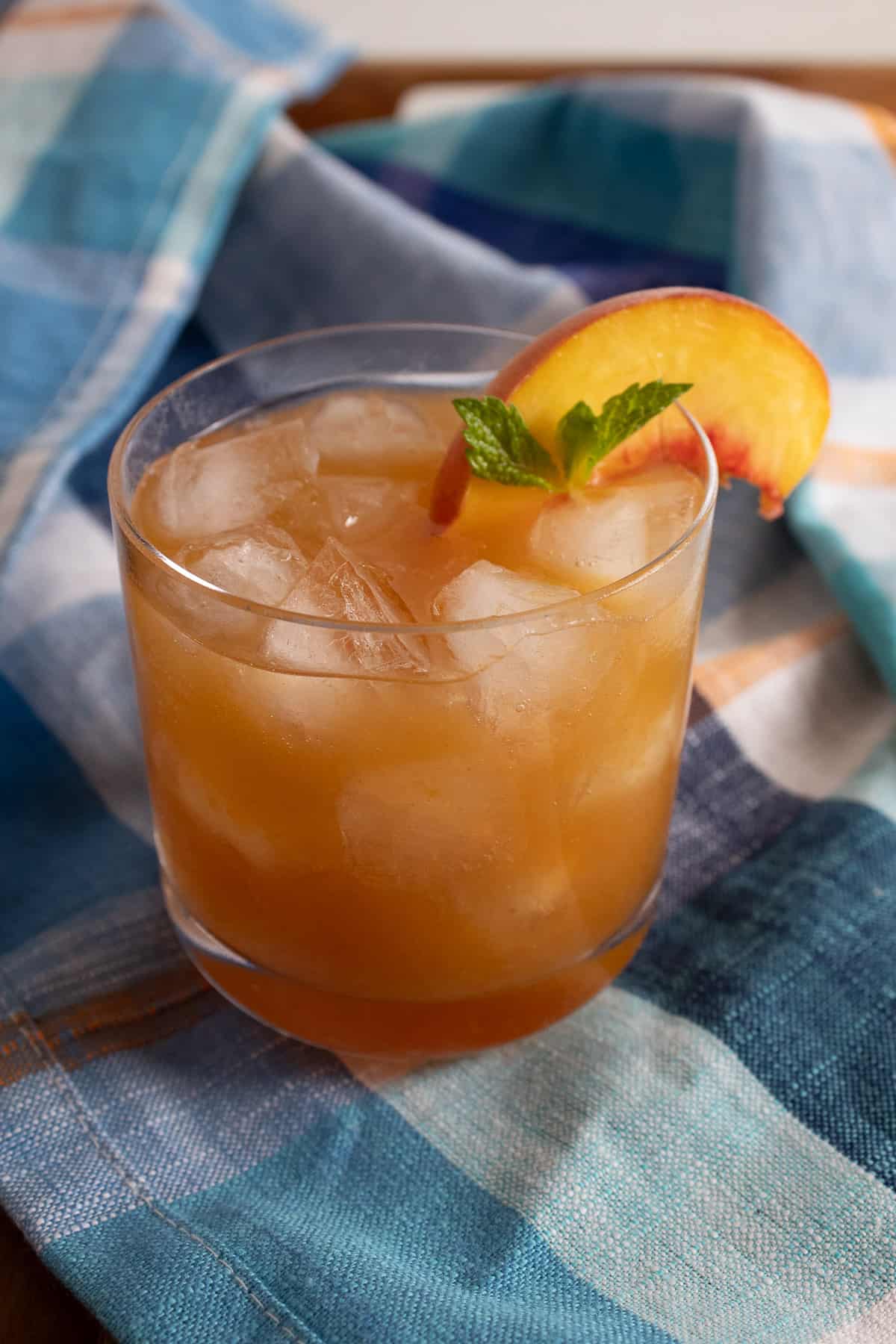 A golden brown beverage is displayed in a short rocks glass and garnished with a peach slice and small mint leaves.