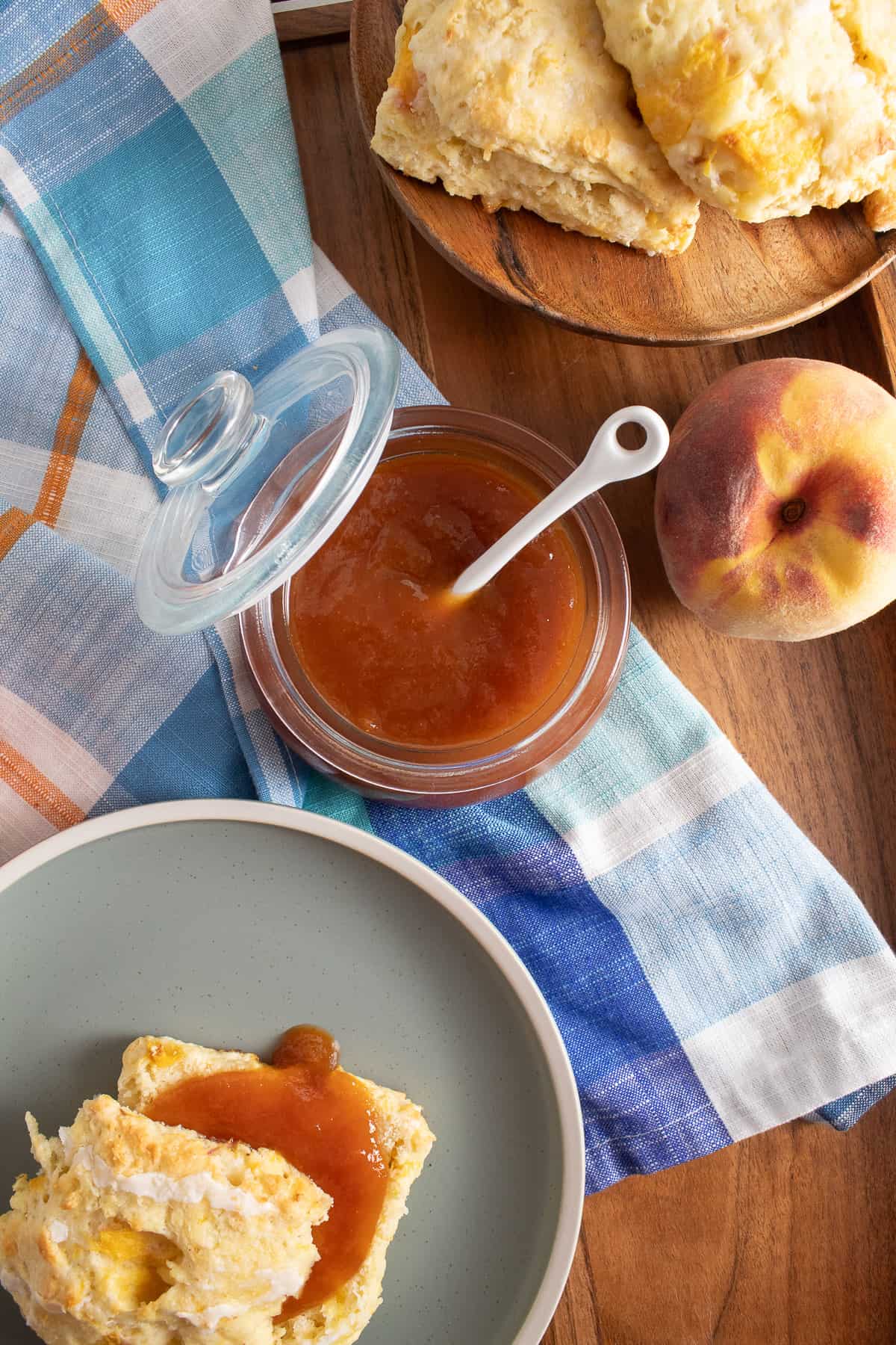 A jar of peach butter sits on a wood tray along with plates of biscuits, a fresh peach, and a blue plaid cloth.