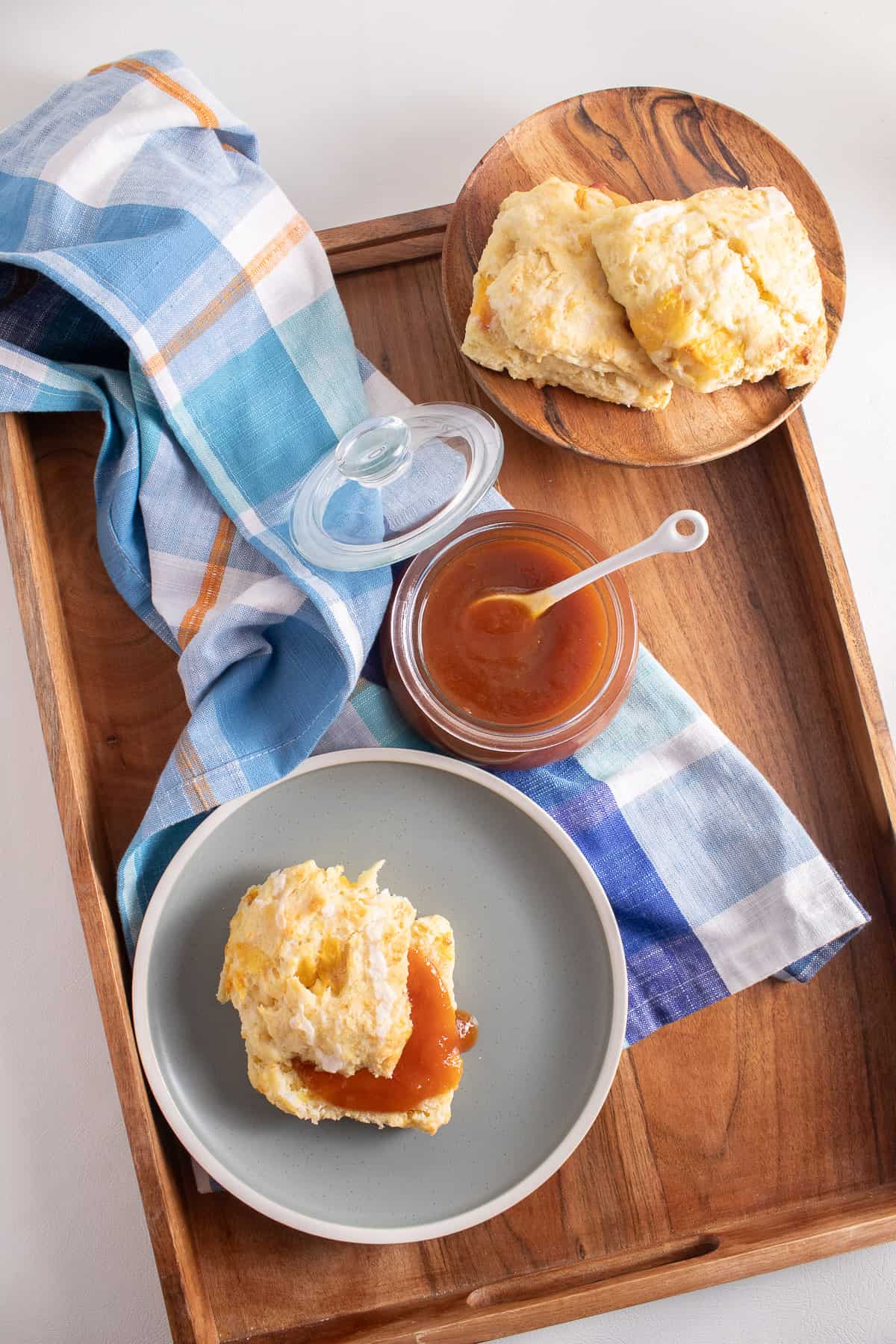 A jar of peach butter sits on a wood tray along with plates of biscuits and a blue plaid cloth.