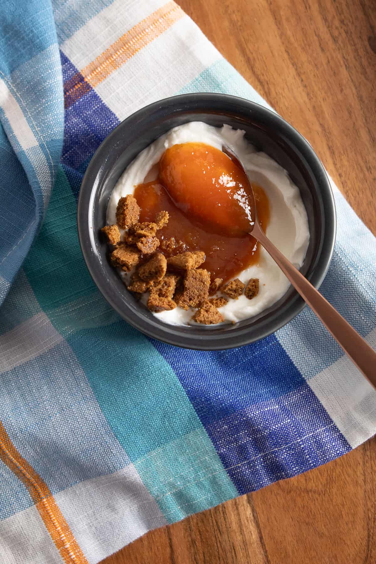 A gray bowl is filled with yogurt, peach butter, and crushed gingersnaps and sits on a blue plaid cloth.