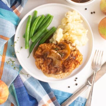 A serving of golden breaded pork and apples on a white plate with gravy, green beans, and mashed potatoes.