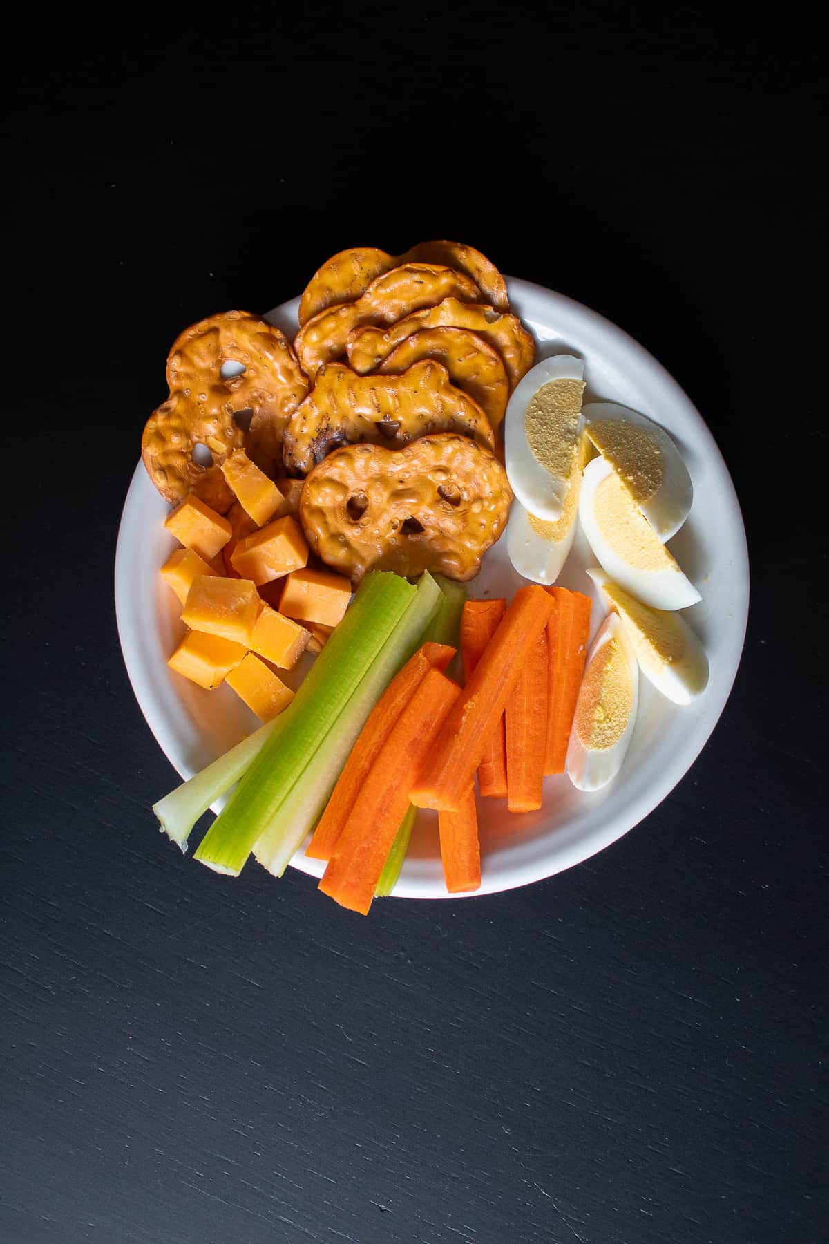 A white plate with hard boiled eggs, pretzels, cheese cubes, and vegetable sticks sits on a black surface.