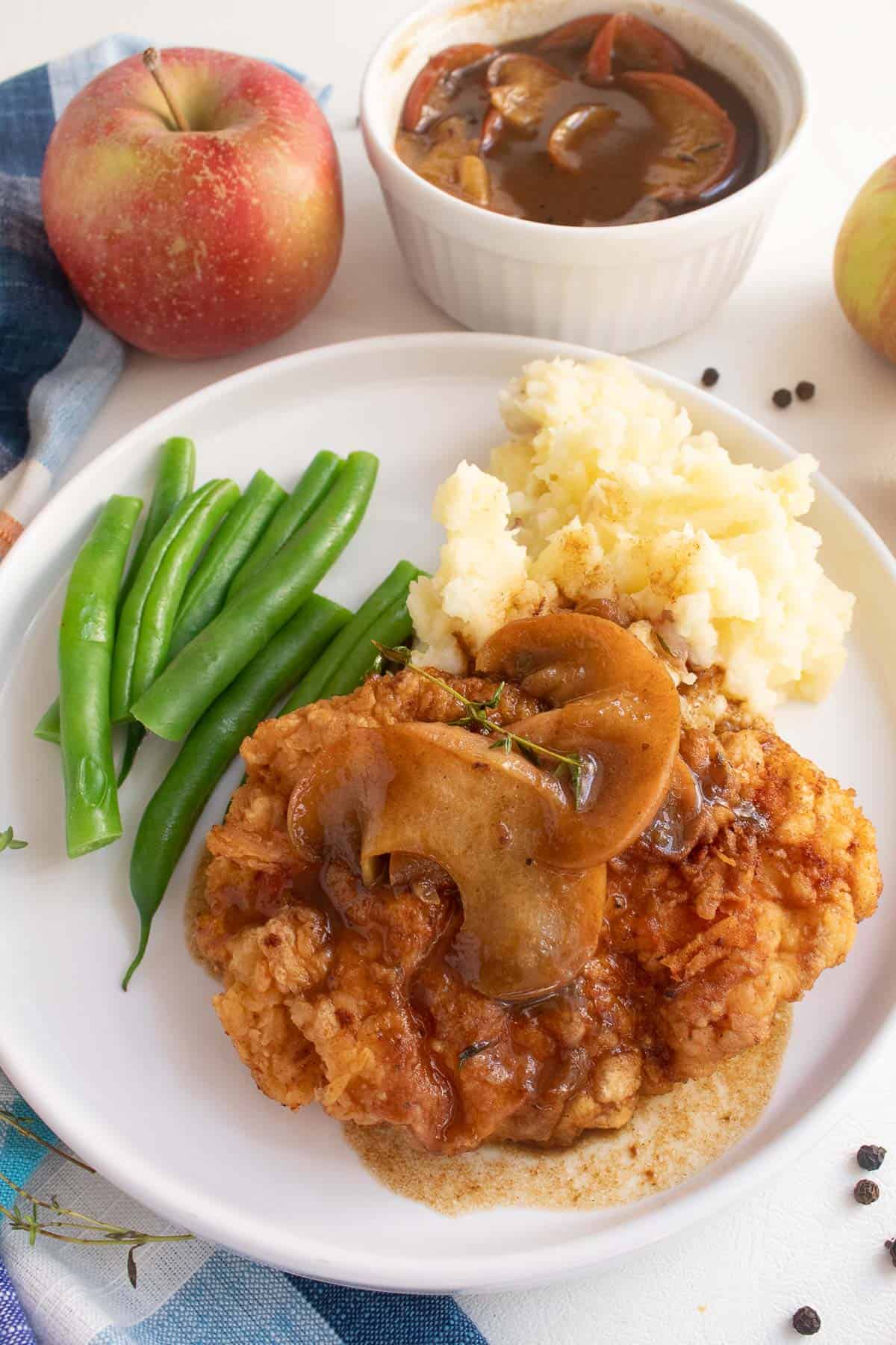 A serving of golden breaded pork and apples on a white plate with gravy, green beans, and mashed potatoes.