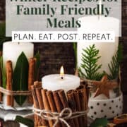 Candles wrapped in cinnamon sticks, bay leaves, and evergreen stems sit on a wooden surface. The words, "20 warming winter recipes for family friendly meals" and "plan. eat. post. repeat." are in two boxes overlaid on the image.