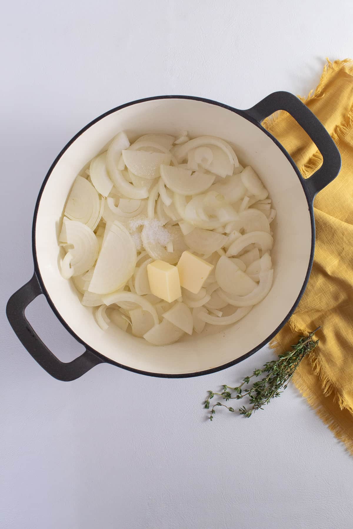 Sliced onions, butter, and salt are combined in a large Dutch oven sitting on a white surface next to a yellow napkin and a thyme sprig.
