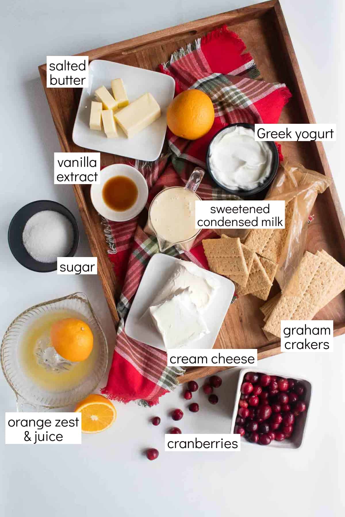 Ingredients for cranberry cheesecake bars with orange cranberry swirl arranged on a wood tray. Text labels in white boxes include: salted butter, vanilla extract, Greek yogurt, sweetened condensed milk, sugar, cream cheese, graham crackers, orange zest & juice, and cranberries.