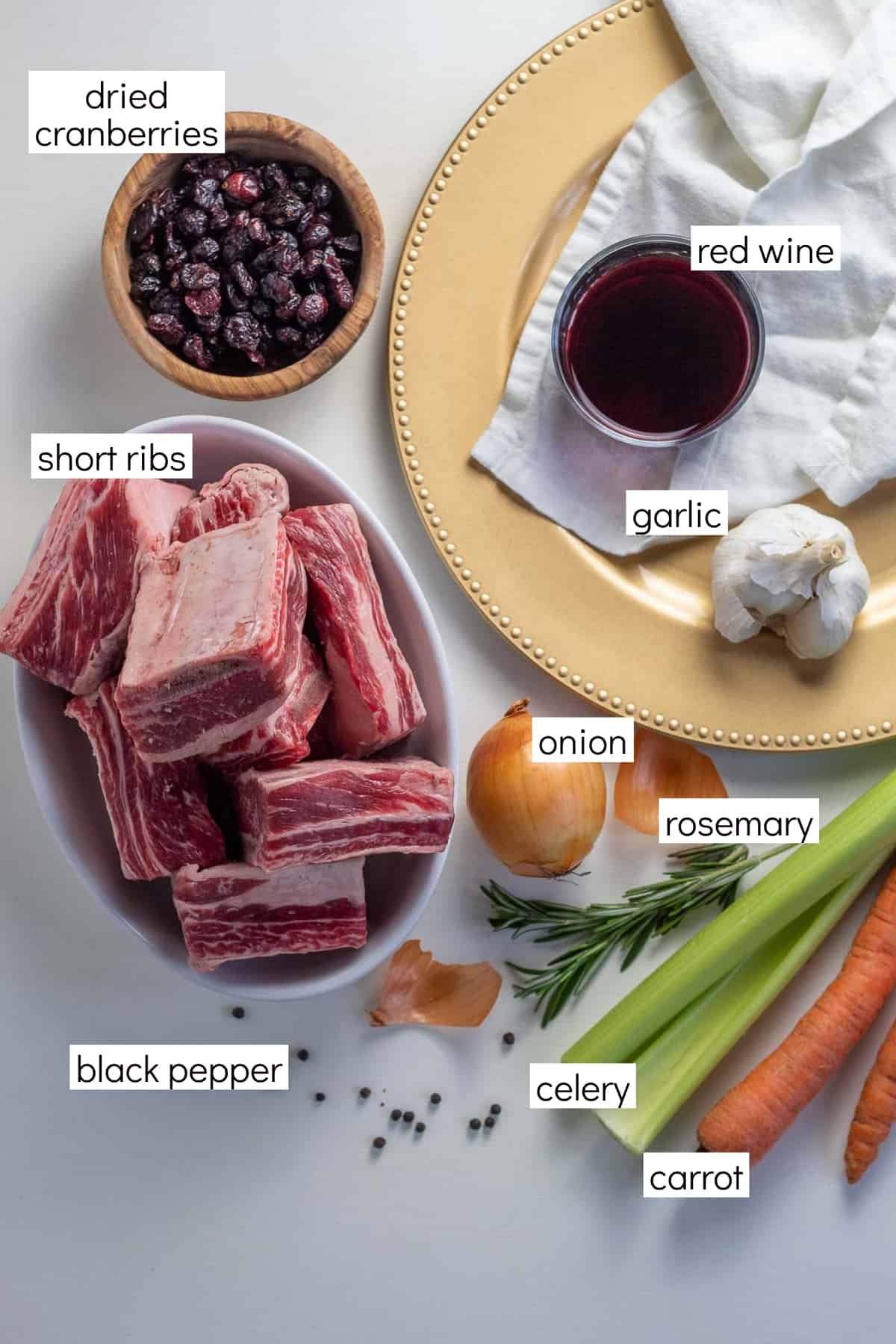 Ingredients for the recipe are displayed on a white surface with labels in white boxes, including: dried cranberries, red wine, short ribs, garlic, onion, rosemary, black pepper, celery, and carrot.