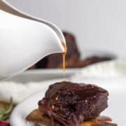 Short rib glaze is poured over a short rib on a white plate from a gravy boat.