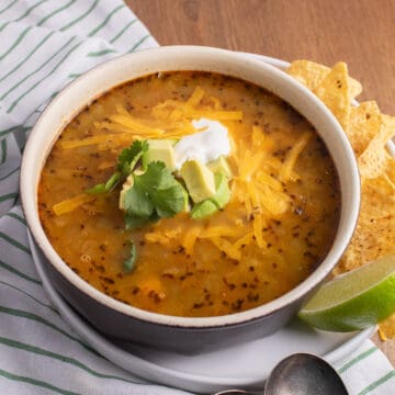 A bowl of the soup is topped with cilantro, avocado chunks, shredded cheese, and sour cream and is accompanied by a handful of tortilla chips and a lime wedge.