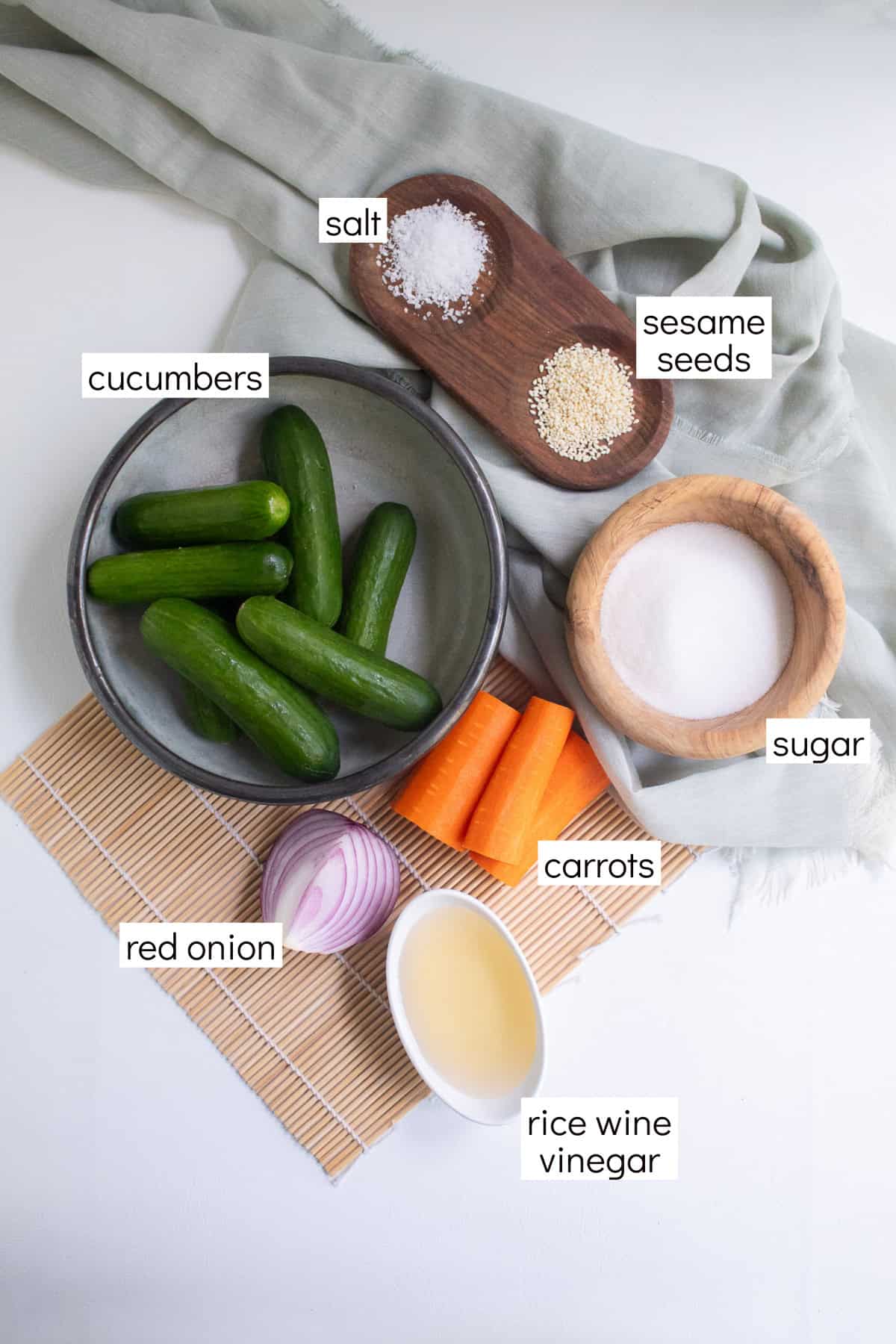 Ingredients for the salad are displayed on a bamboo and green cloth surface. White text boxes label the ingredients, including salt, cucumbers, red onion rice wine vinegar, carrots, sugar, and sesame seeds.