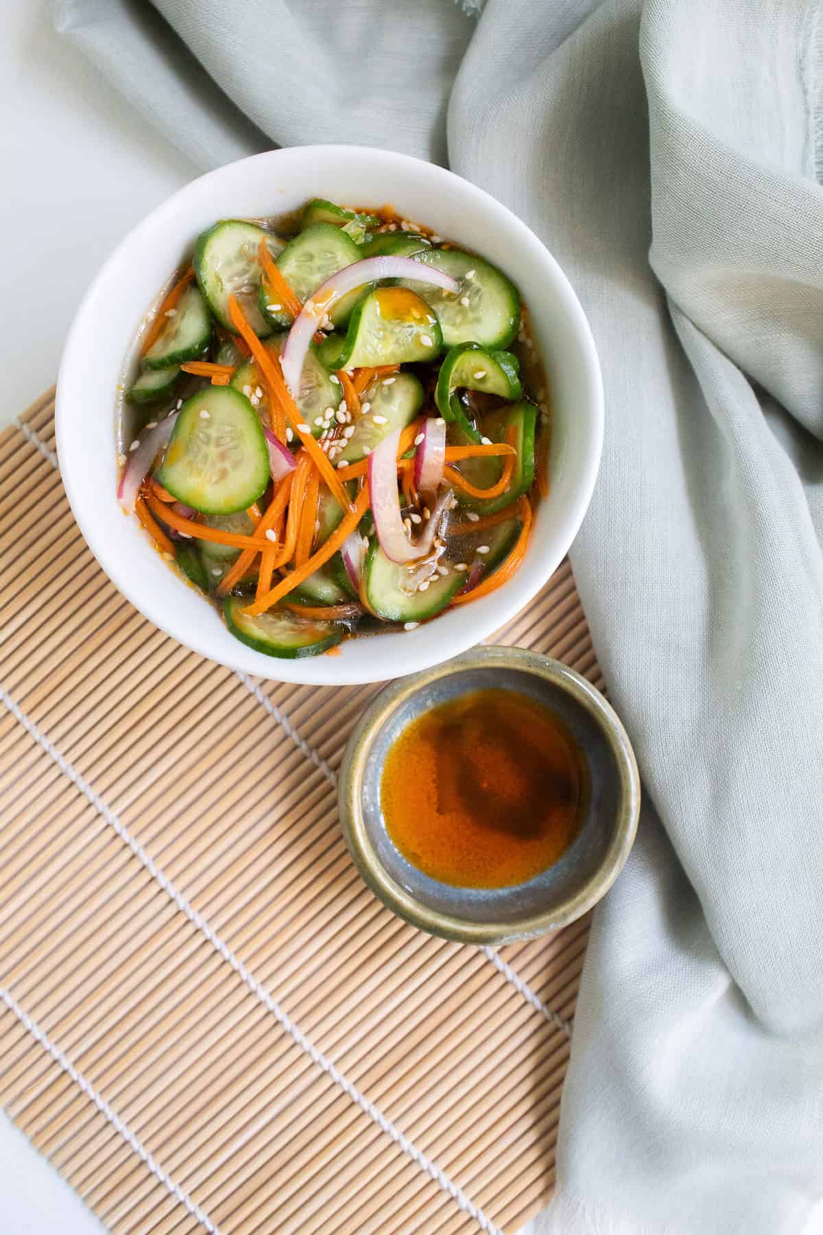A bowl of spicy cucumber salad sits next to a small dish of chili oil on a bamboo mat.